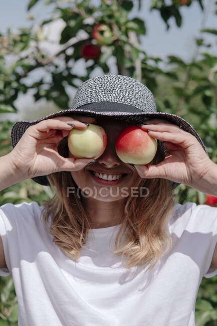 Femme joyeuse portant un chapeau tenant des pommes devant les yeux au verger — Photo de stock
