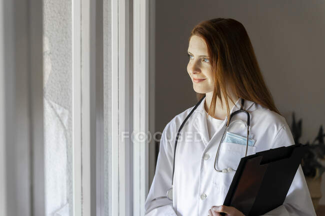 Nachdenkliche junge Ärztin lächelt beim Blick aus dem Fenster im Home Office — Stockfoto