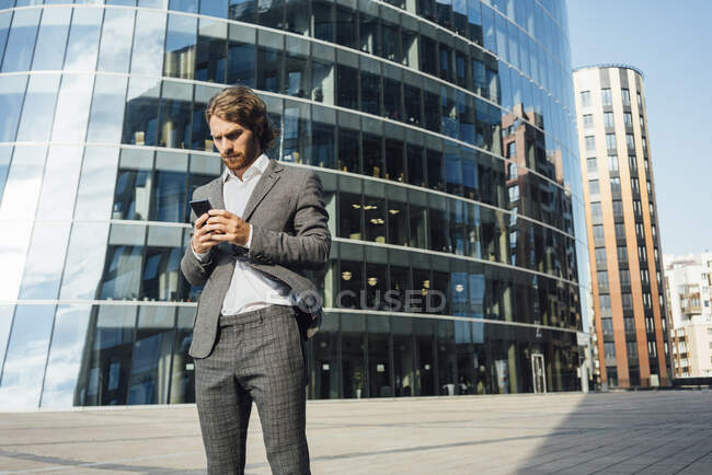 Handsome young businessman using smart phone while standing against modern office building in downtown on sunny day — Stock Photo