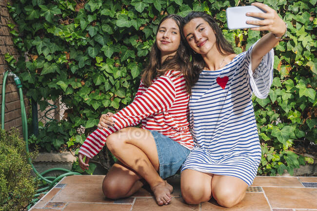 Sorelline sorridenti mentre scattano selfie sullo smart phone mentre si inginocchiano nel cortile — Foto stock
