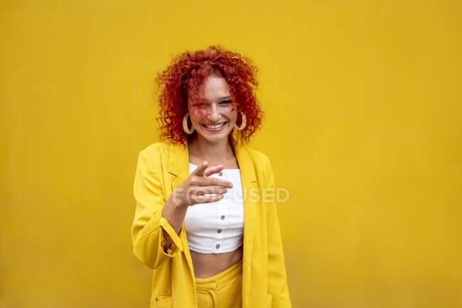 Happy young woman with red curly hair pointing with finger in front of yellow wall — Foto stock