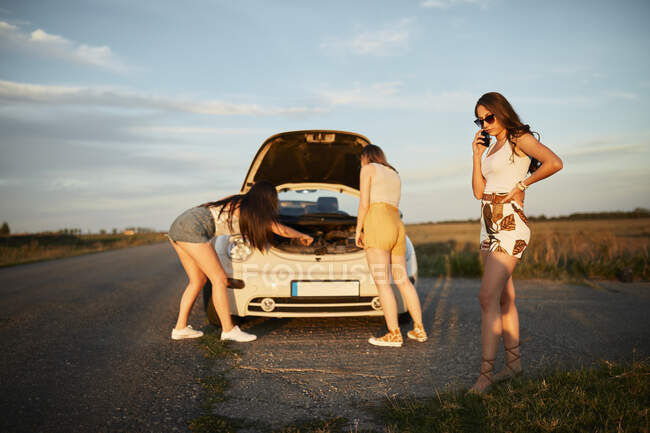 Жінка розмовляє по мобільному телефону, а друзі ремонтують машину по дорозі на заході сонця. — стокове фото