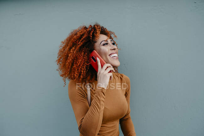 Joyeux jeune femme avec des cheveux afro parler sur téléphone portable tout en se tenant contre le mur — Photo de stock