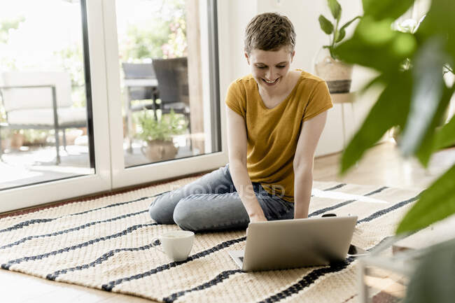 Smiling mid adult woman with short hair using laptop while sitting on carpet at home — Fotografia de Stock