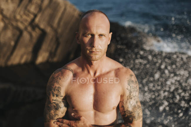 Tattooed nudist standing on volcanic rocks by the sea with arms crossed â€”  one person, 30 40 Years - Stock Photo | #479997000