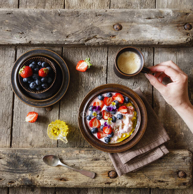 Женская рука тянется за чашкой кофе, стоящей рядом с тарелкой кварка с фруктами и съедобными цветами — стоковое фото
