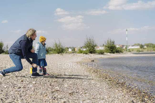 Pai e filha olhando para o rio enquanto joga seixos na água perto da margem do rio — Fotografia de Stock