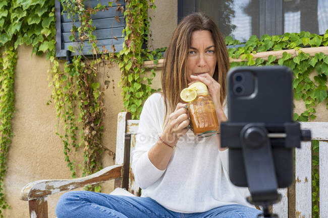 Женщина-диетолог пьет сок из банки во время видеоблога через смартфон на заднем дворе — стоковое фото