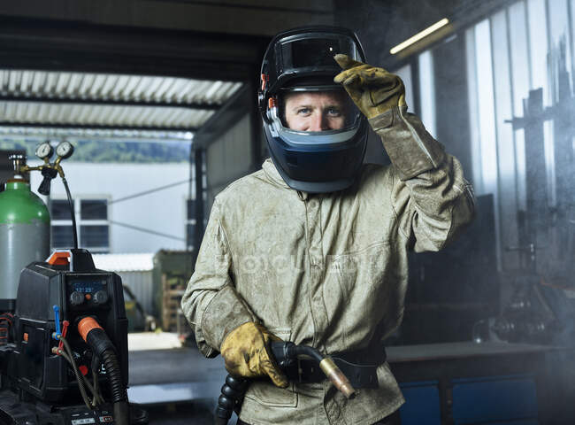 Manual worker wearing protective welding helmet holding welding machine while standing at factory — Stock Photo