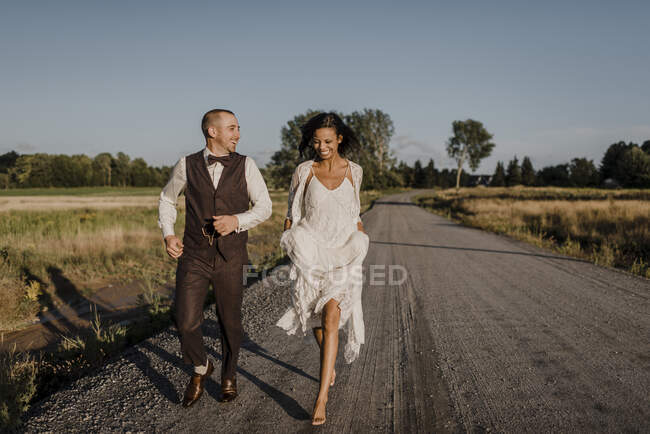 Noiva feliz e noivo correndo na estrada durante o dia ensolarado — Fotografia de Stock
