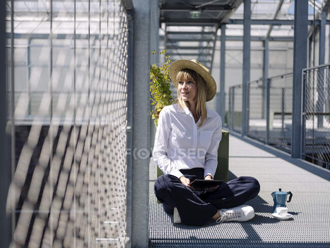 Деловая женщина в шляпе с помощью цифрового планшета, сидя у забора в теплице — стоковое фото