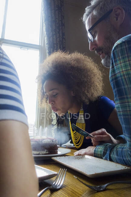 Young woman wearing ethnic jewelry blowing candles on cake while sitting with friends at cafe — Stock Photo