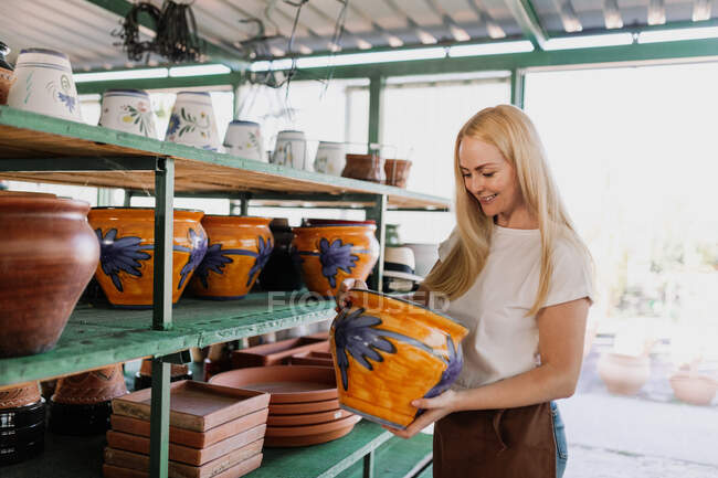 Dueña sonriente sosteniendo cerámica mientras está de pie junto al estante en el taller - foto de stock