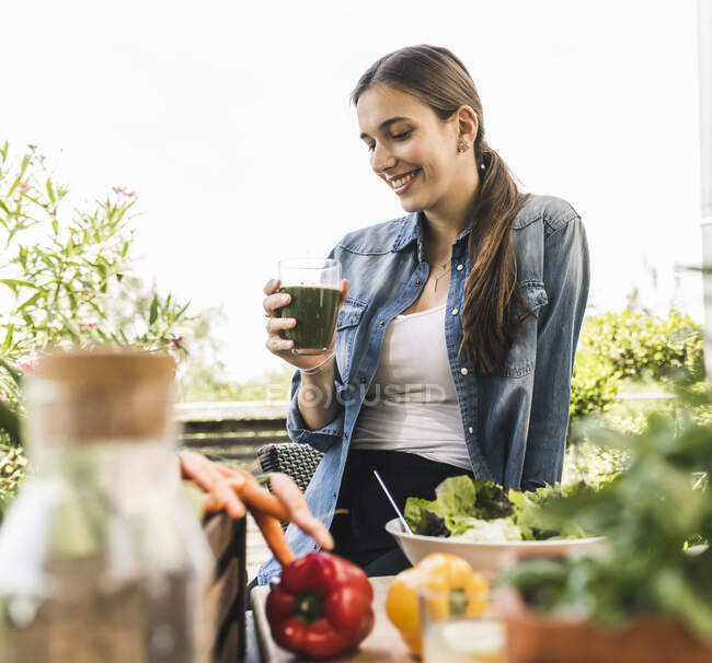 Mujer sonriente sosteniendo jugo mientras se sienta contra el cielo despejado en el patio - foto de stock