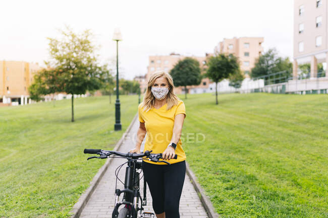 Mujer con mascarilla protectora de pie con bicicleta en el sendero de la ciudad durante COVID-19 - foto de stock