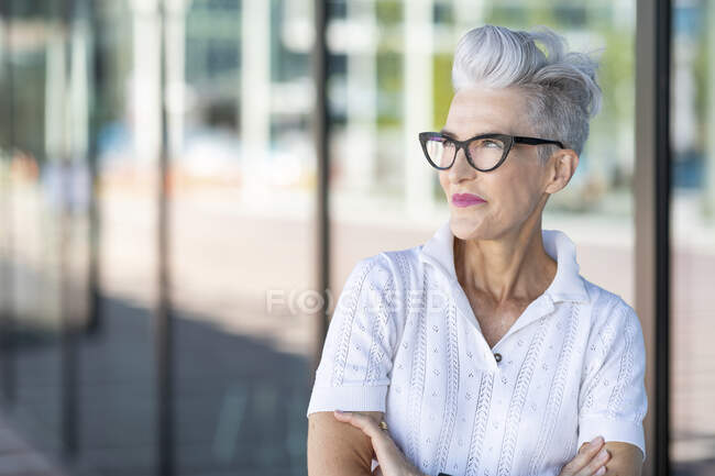 Smiling senior woman with arms crossed looking away while standing against glass window — Foto stock