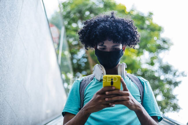 Young man with wearing protective face mask text messaging on smart phone while standing on escalator — Stock Photo