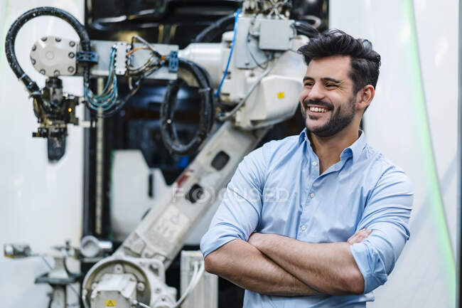 Sorridente imprenditore maschio con le braccia incrociate guardando altrove mentre in piedi da braccio robotico all'industria — Foto stock