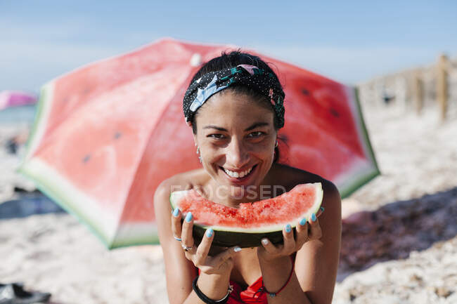 Primer plano de la mujer sonriente sosteniendo la sandía mientras se sienta contra el paraguas rojo en la playa - foto de stock