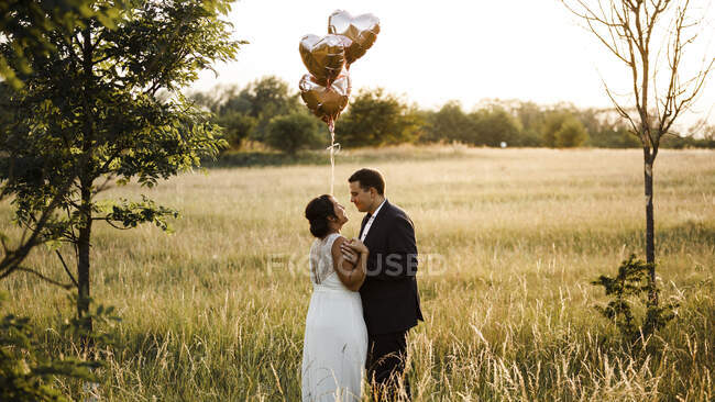 Brautpaar hält herzförmige Heliumballons, während es bei Sonnenuntergang auf dem Feld steht — Stockfoto