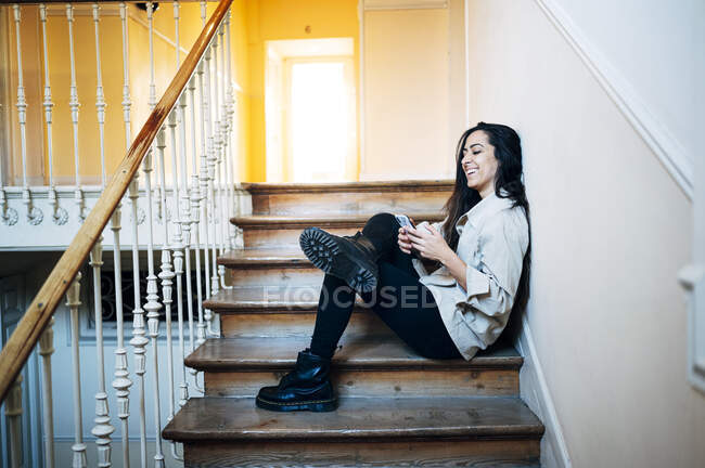 Mujer joven feliz usando el teléfono inteligente mientras está sentado en la escalera en casa - foto de stock