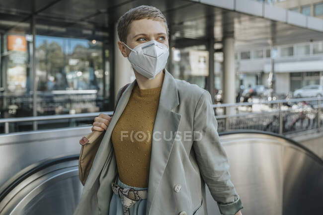 Женщина в защитной маске смотрит в сторону, взбираясь на эскалатор в городе — стоковое фото