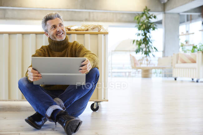 Uomo sorridente con tablet digitale che distoglie lo sguardo mentre è seduto sul pavimento a casa — Foto stock