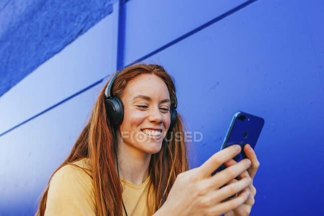 Рыжая женщина в наушниках с помощью мобильного телефона, сидя у синей стены — стоковое фото