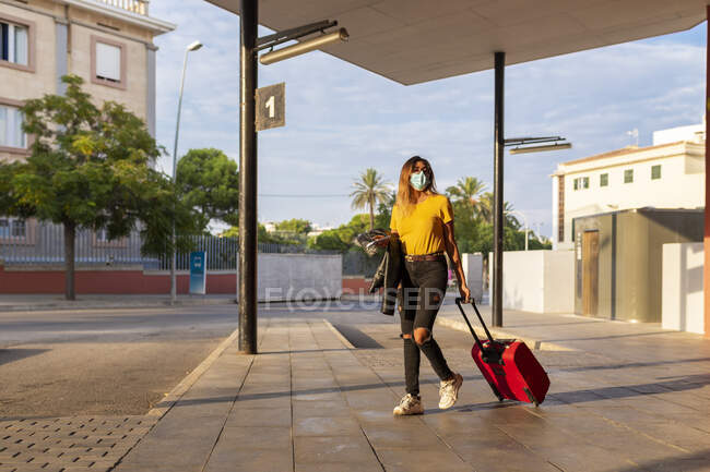 Mujer joven con máscara protectora mientras camina con equipaje en la parada de autobús - foto de stock