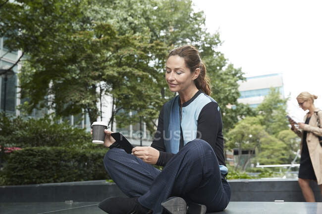 Femme d'affaires tenant une tasse de café tout en utilisant un téléphone portable avec un collègue debout en arrière-plan au parc de bureau — Photo de stock