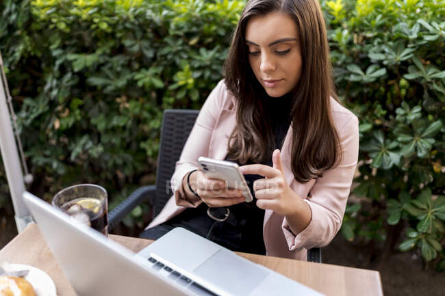 Joven empresaria usando teléfono inteligente mientras está sentada en la terraza de la cafetería - foto de stock