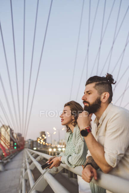 Couple looking away while leaning on railing at bridge in city during sunset — Stock Photo