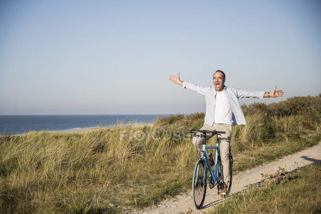 Heureux homme mature avec les bras tendus vélo d'équitation à la plage contre le ciel clair — Photo de stock