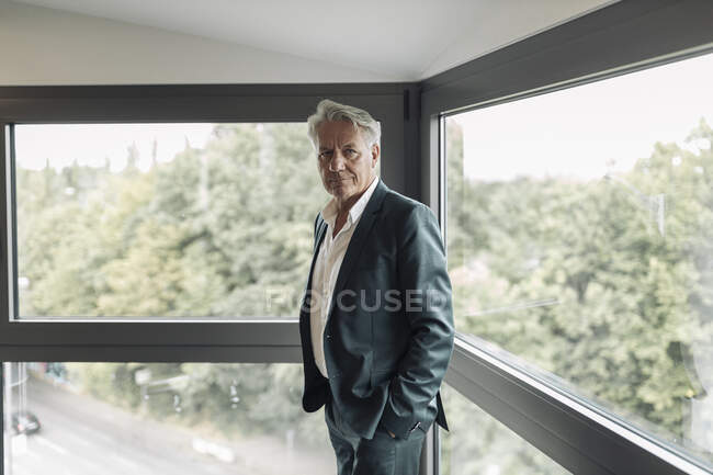 Empresario con la mano en bolsillos de pie junto a la ventana en la oficina - foto de stock