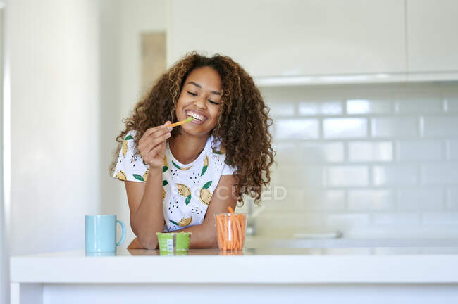 Sorrindo jovem mulher tendo guacamole com cenouras na cozinha — Fotografia de Stock