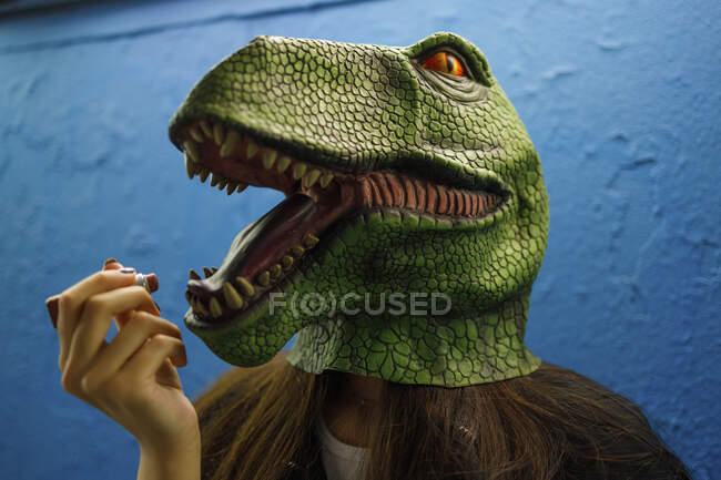 Mujer aplicando lápiz labial mientras usa máscara de dinosaurio contra la pared azul - foto de stock