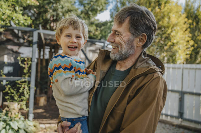Père riant en portant son fils debout dans la cour arrière par une journée ensoleillée — Photo de stock