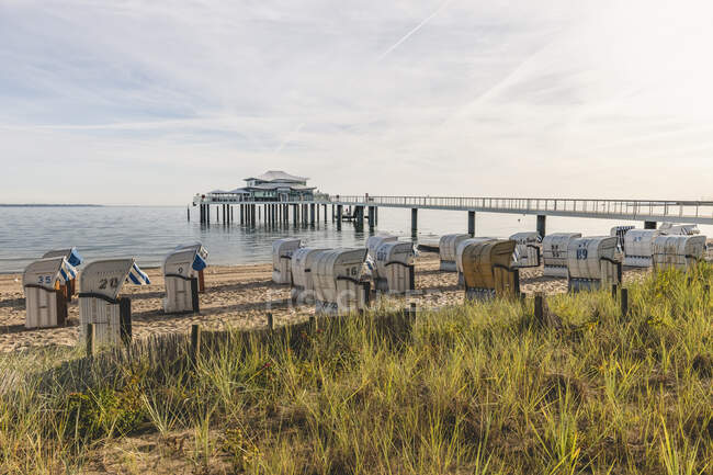 Alemania, Schleswig-Holstein, Timmendorfer Strand, Sillas de playa con capucha en la playa costera de arena con casa de té al final del muelle en el fondo - foto de stock