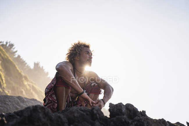 Jeune homme regardant loin tout en s'accroupissant sur le rocher à la plage contre le ciel — Photo de stock