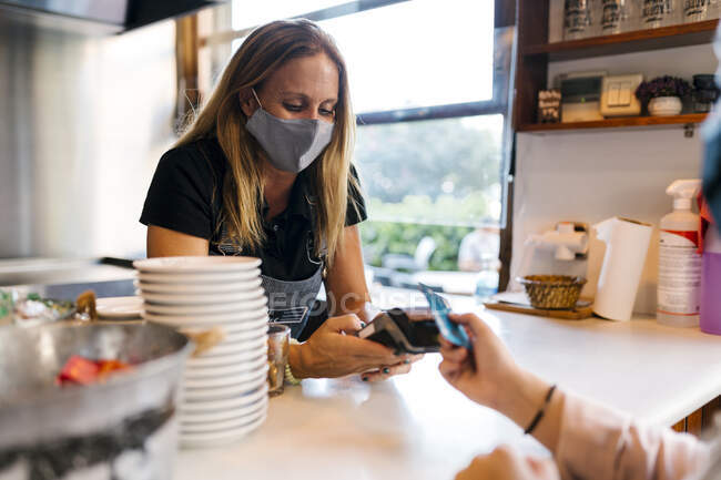 Жіночий касир в масці обличчя отримує оплату через кредитну картку в кафе під час COVID-19. — стокове фото