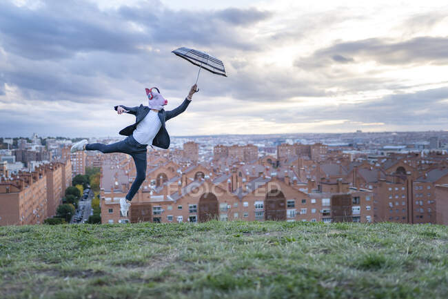 Male professional wearing pig mask holding umbrella while jumping on hill at sunset — Foto stock