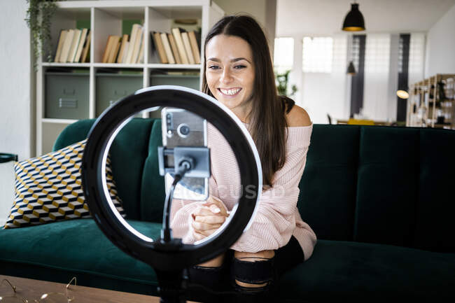 Sonriendo hermosa mujer influencer sentado en el sofá mientras vlogging a través de teléfono inteligente en casa - foto de stock