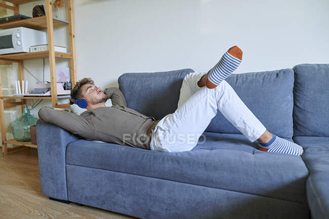 Hombre joven con auriculares escuchando música mientras está acostado en el sofá en casa - foto de stock