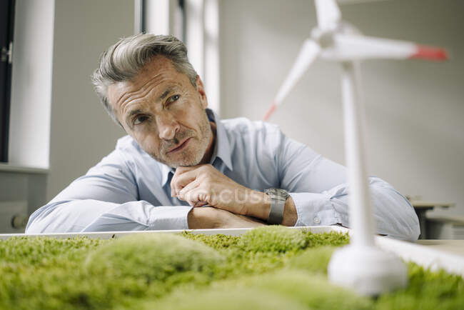 Empresário olhando para o brinquedo da turbina eólica sobre o quadro de musgo enquanto se inclina na mesa no escritório — Fotografia de Stock