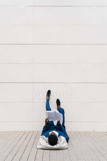 Молодий бізнесмен лежить з мішком для сміття на тротуарі, читаючи книжку в місті. — стокове фото