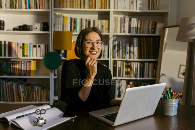 Femme d'affaires avec la main sur le menton en utilisant un ordinateur portable tout en étant assis contre la bibliothèque — Photo de stock