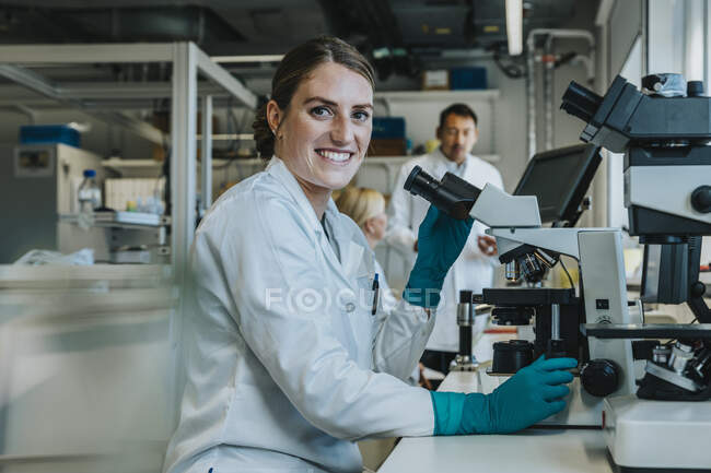 Smiling woman analyzing human brain slide under microscope while sitting with scientists in background at laboratory — Stock Photo