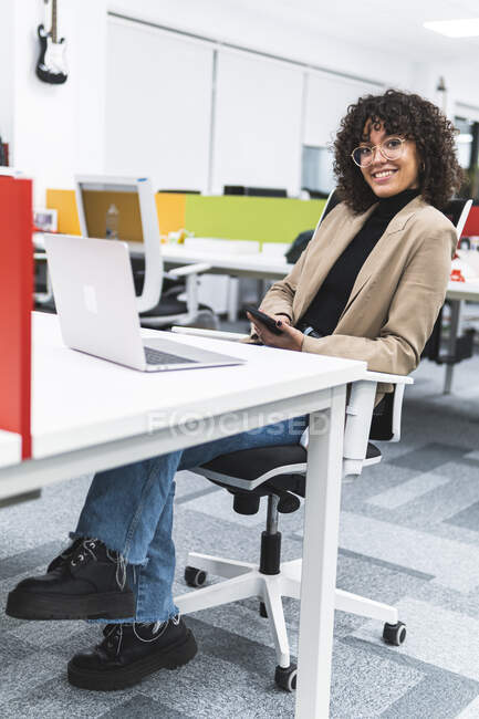 Professionista sorridente con computer portatile che tiene il telefono cellulare mentre è seduto sulla scrivania in ufficio — Foto stock