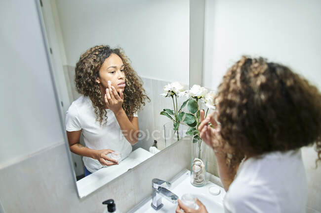 Hermosa mujer que aplica crema facial mientras mira el reflejo del espejo en el baño - foto de stock