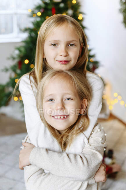 Smiling sisters embracing with Christmas tree in background at home ...